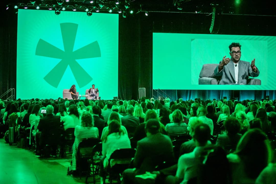 A large audience is seated in a conference hall with bright green lighting. On stage, two speakers are engaged in discussion, with one speaker animatedly gesturing while seated on a grey couch. Behind them, a large screen displays a prominent green asterisk logo, creating a vibrant backdrop for the presentation.