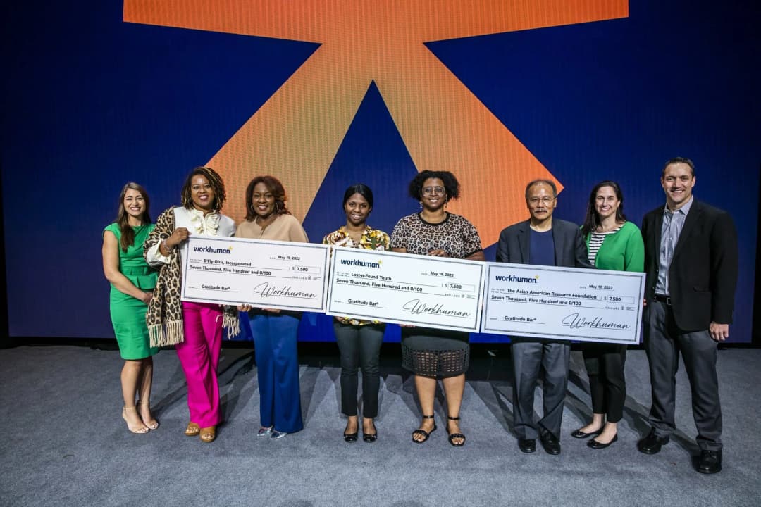 A group of individuals stands together on stage, each holding oversized checks from Workhuman, which are meant for various organizations: B'Fly Girls, Incorporated, Lost-n-Found Youth, and The Asian American Resource Foundation, each check for $7,500. The background features an orange graphic design on a blue backdrop. The people are smiling and appear to be celebrating the moment.