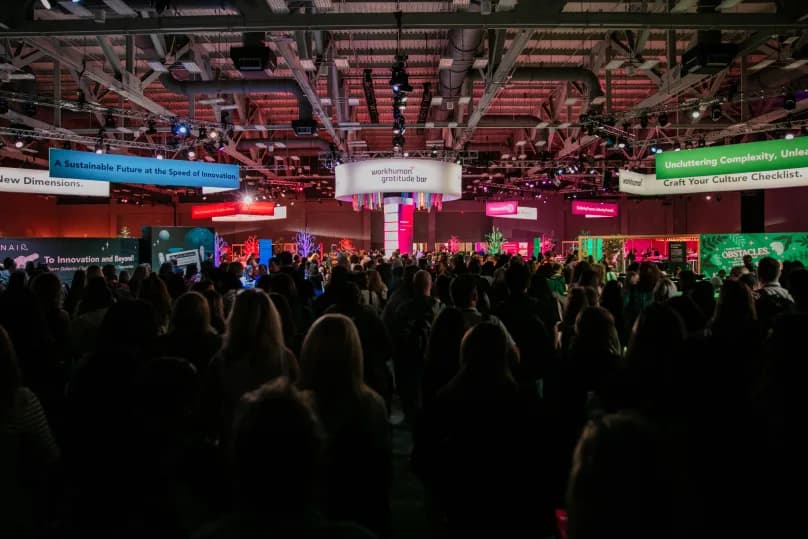 A large crowd is gathered at a Workhuman event, engaged in activities around colorful displays and signage. Prominent elements include a 'Workhuman Gratitude Bar' and banners highlighting themes such as 'A Sustainable Future at the Speed of Innovation' and 'Uncluttering Complexity.' The scene is lively with various lighting and decorations creating a vibrant atmosphere.