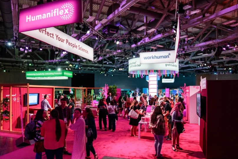 A vibrant Workhuman event space featuring a bustling crowd interacting in front of various displays. Key elements include a large sign with 'Humaniflex' and 'Perfect Your People Science,' along with a central 'workhuman gratitude bar' sign. The area is filled with colorful decorations and a lively atmosphere as attendees engage with exhibits and each other.