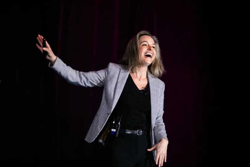 A woman smiling and enthusiastically gesturing with her hand while speaking to an audience. She is wearing a black top and a patterned blazer, standing against a dark backdrop.