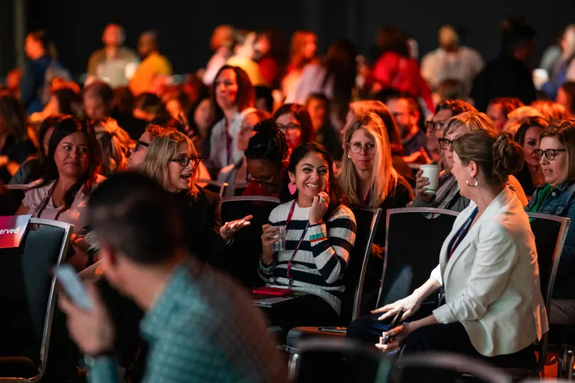 A lively audience at a Workhuman event, with a diverse group of attendees chatting and enjoying the atmosphere. A woman in a striped shirt and pink earrings is smiling and engaging with her surroundings, while others are seen laughing, holding cups, and sitting in rows of chairs. The overall mood is energetic and collaborative, capturing a moment of connection among participants.