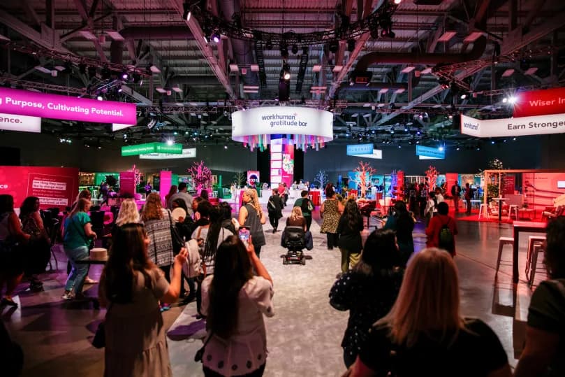 A large conference space filled with attendees walking towards a central display labeled 'workhuman gratitude bar.' The setting features brightly colored signage and decorations, with various sections highlighted in pink, green, and blue. People are engaging with the space, and several trees and plants add to the vibrant atmosphere.
