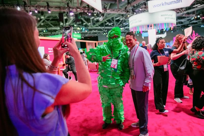 A group of attendees at a Workhuman event, with one person dressed in a bright green costume resembling a caricature made of paper strips, posing for a photo with another attendee. In the background, various event signage and other attendees are visible, all set against a vibrant pink carpet. The atmosphere appears lively and engaging.