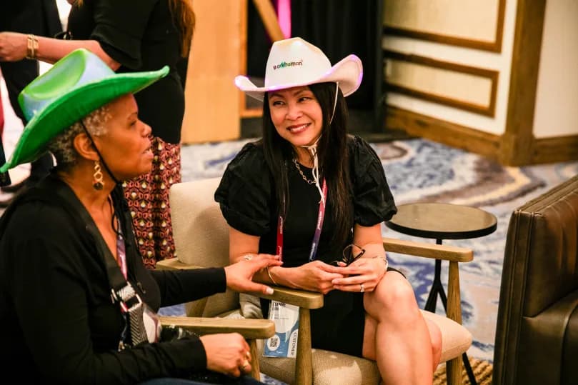 Two women are sitting in a casual setting, engaging in conversation. One woman, wearing a green cowboy hat, has her hands resting on a chair as she talks. The other woman is seated, wearing a white cowboy hat with the Workhuman logo, and is smiling while listening. The background suggests a relaxed environment, possibly at a conference.