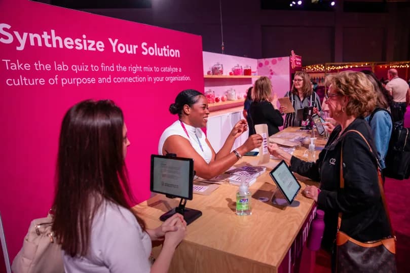 A vibrant conference display featuring a booth labeled 'Synthesize Your Solution,' with attendees engaged in conversation. A sign promotes taking a lab quiz intended to help foster a culture of purpose and connection within organizations. One person, smiling, is handing out materials while others, including a woman with short hair and a white top, are engaged in discussion. Tablets are visible on the wooden table, and the background features additional attendees involved in various activities.