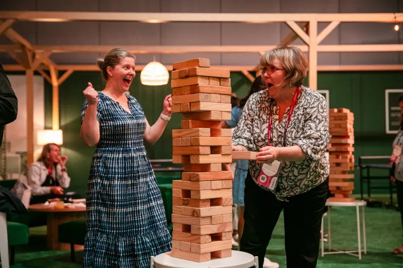 Two women are joyfully interacting in a lively setting while playing Jenga. One woman, wearing a blue striped dress, raises her arm in excitement, while the other, in a patterned shirt, appears shocked as they witness a tall stack of wooden blocks about to topple. The background features comfortable seating and people engaged in conversation, adding to the event's cheerful atmosphere.