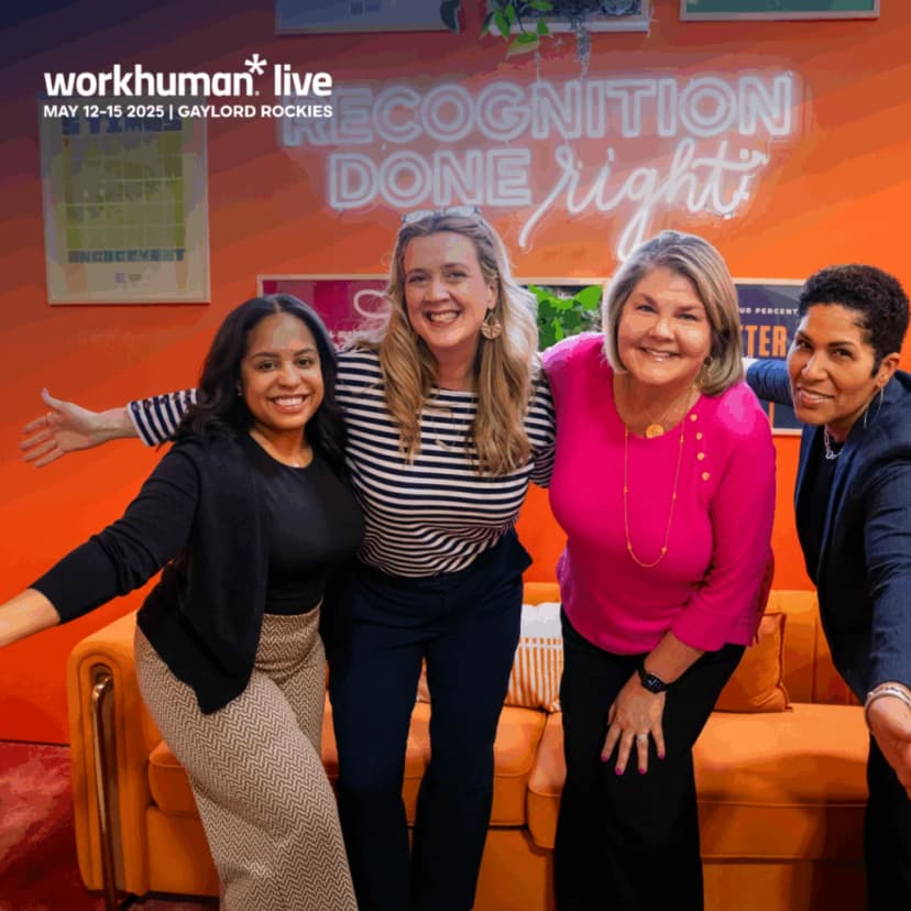 A group of four women posing together in front of an orange backdrop featuring the text 'RECOGNITION DONE right.' They are smiling and appear to be celebrating, with one woman wearing a striped shirt and three others in solid colors. The setting includes a cozy orange couch, hinting at a lively and positive atmosphere.
