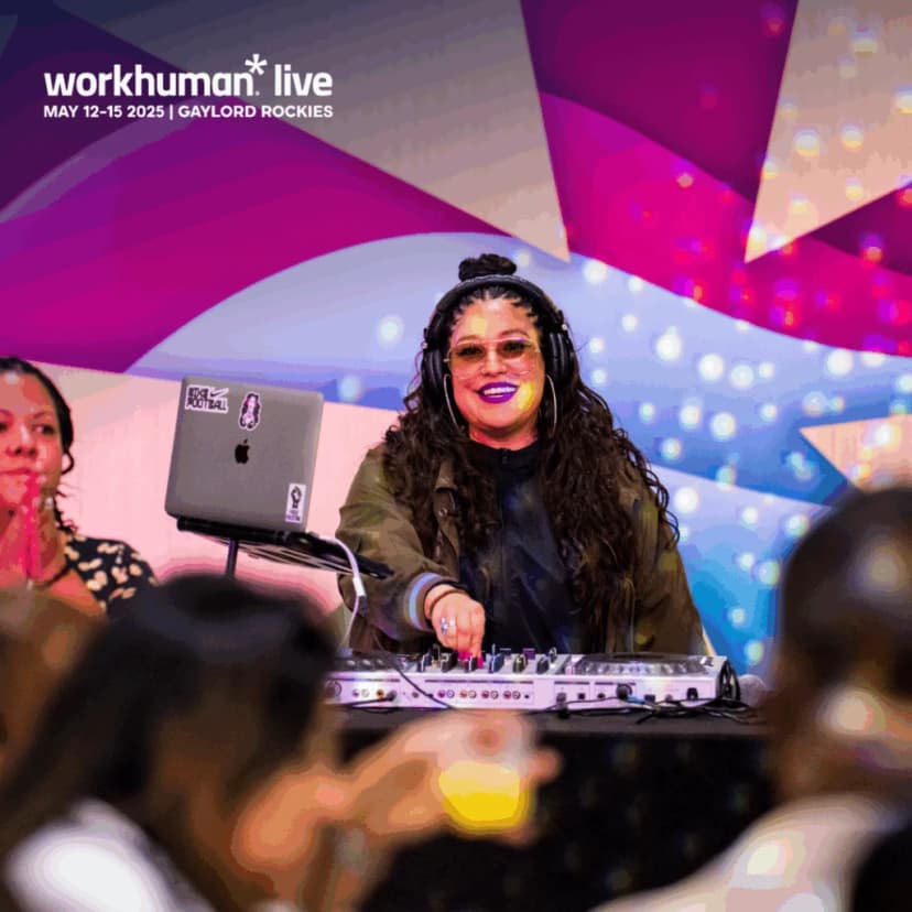 A DJ with long, curly hair and sunglasses performs at a lively event, with a colorful backdrop featuring pink and blue swirls. In the foreground, attendees are seen enjoying the atmosphere.