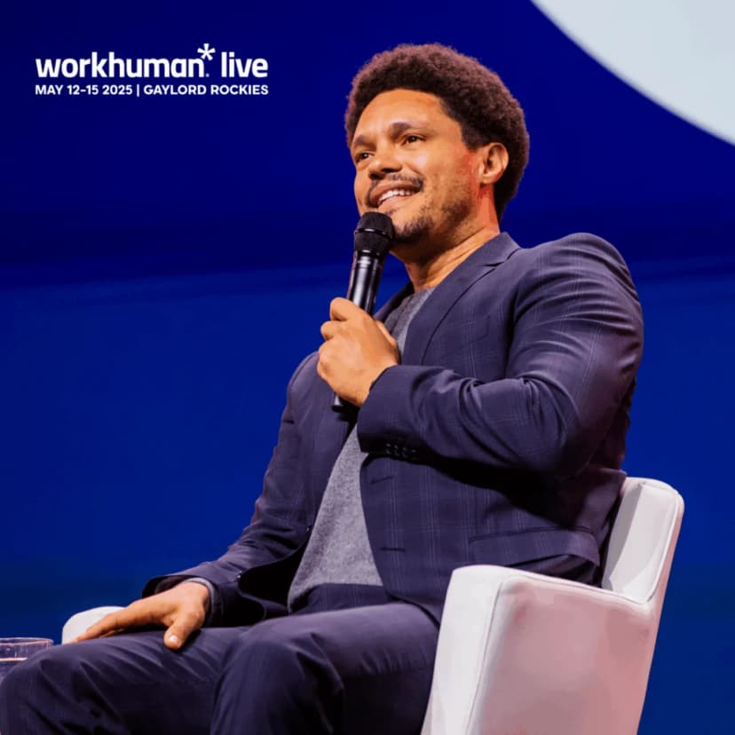 A man with curly hair sits on a white chair, holding a microphone and smiling while speaking. He is dressed in a navy blazer over a grey shirt. The background features a blue gradient.