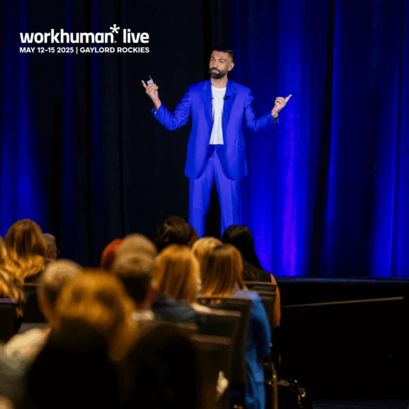 A person in a bright blue suit stands on stage, gesturing with their hands while speaking to an audience. The background features dark curtains and a spotlight effect, and the audience is partially visible, showing heads and shoulders.