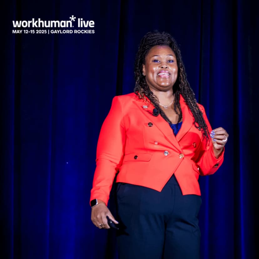 A woman wearing a bright red blazer is speaking at an event, smiling and gesturing with her right hand. The background is dark, emphasizing her presence on stage.