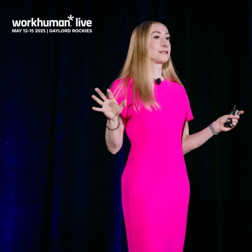 A woman in a bright pink dress is speaking on stage at a conference, with her arms raised in a gesture of emphasis. The background is dark, creating a contrast that highlights her presence.