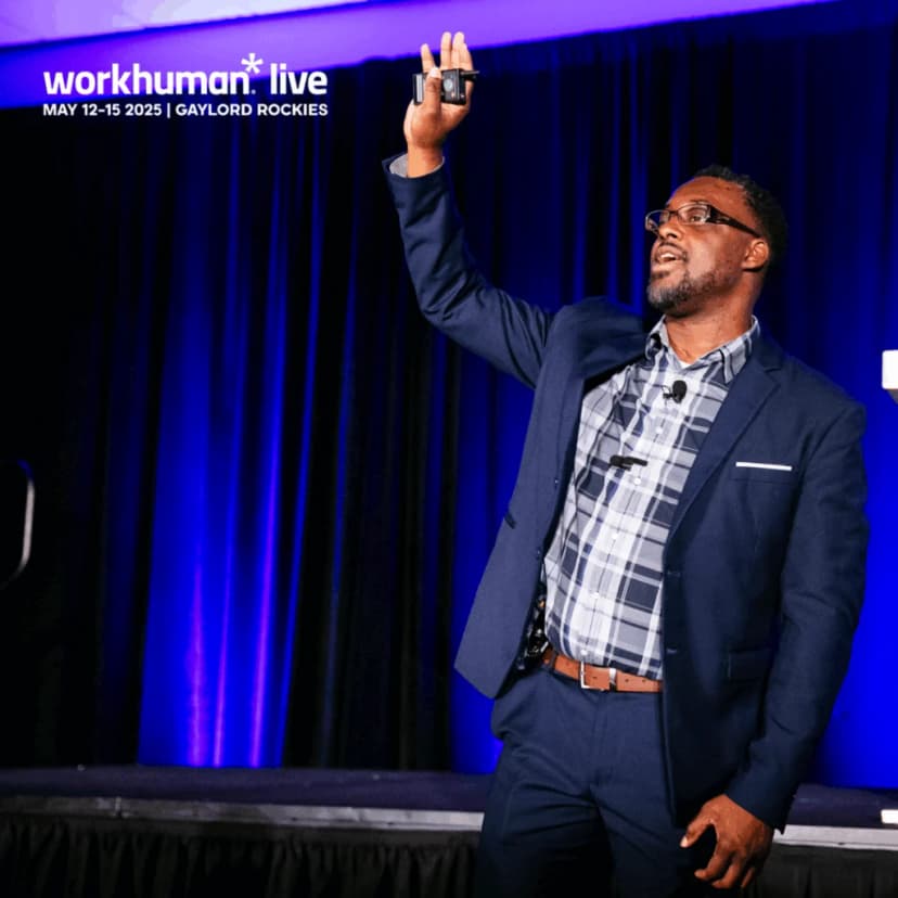 A speaker gestures while presenting at a Workhuman Live event, with the backdrop of a dark curtain illuminated by blue lighting. The speaker is dressed in a plaid shirt and suit, holding a presentation remote in their right hand, and appears engaged with the audience.