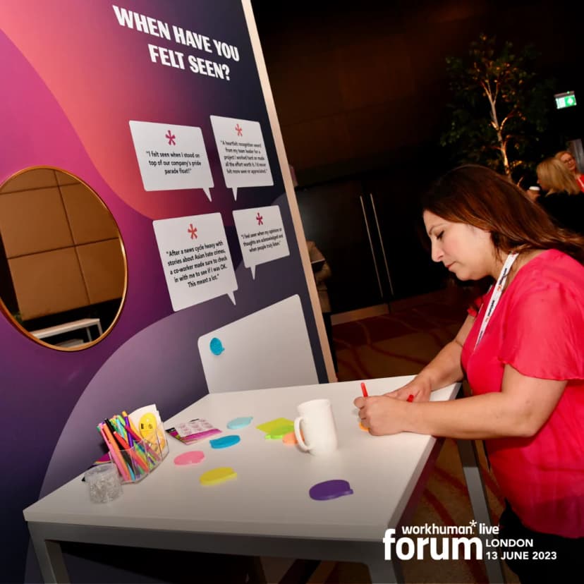 A woman is writing on a table covered with colorful sticky notes at a Workhuman Live forum. Behind her is a large display with the question 'When have you felt seen?' and various recognition quotes related to feeling appreciated and acknowledged. The setting appears to be an interactive space within a conference, with soft lighting and attendees in the background.