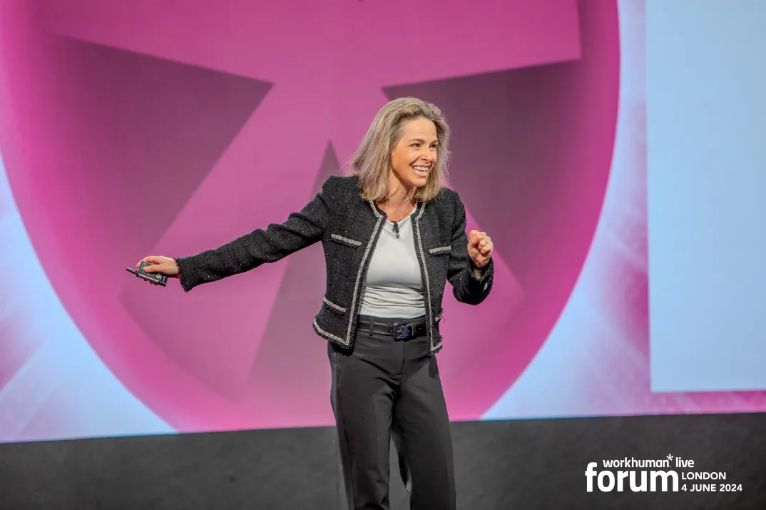 A smiling woman gestures enthusiastically while speaking on stage at the Workhuman Live Forum in London. She is dressed in a dark jacket over a light shirt and stands against a backdrop featuring vibrant geometric shapes.