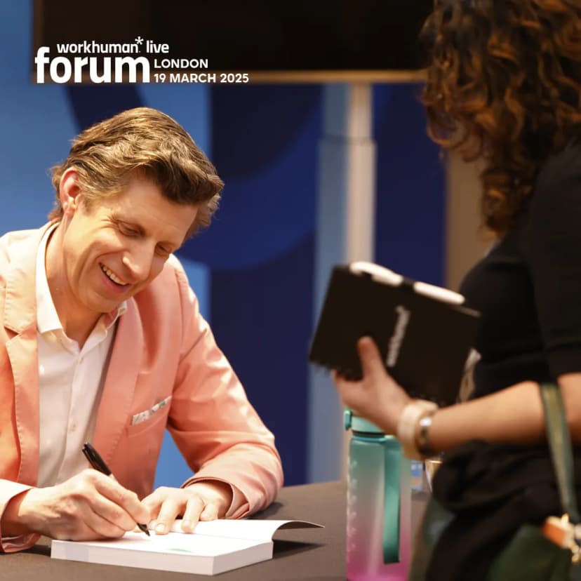 A smiling man in a pink blazer is signing a book at a Workhuman Live event. He is seated at a table, focused on the book, while a woman standing in front of him holds another book.
