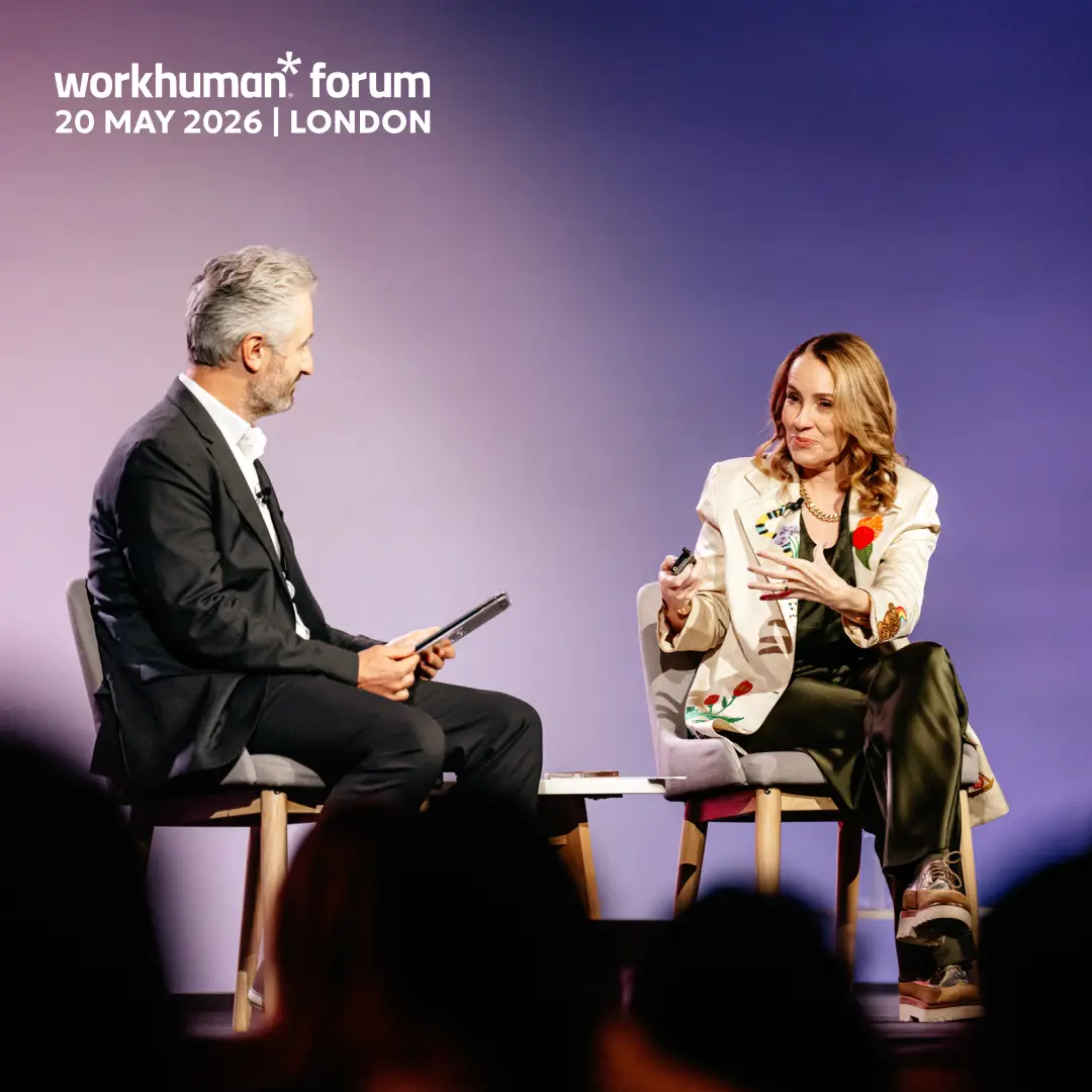 A man and a woman are seated on stage at the Workhuman Forum event in London, scheduled for May 20, 2026. The man, dressed in a black suit with a white shirt, holds a tablet while looking at the woman, who is wearing a stylish, embroidered blazer with green pants. She gestures animatedly as they engage in conversation. The background features a gradient that transitions from purple to blue.