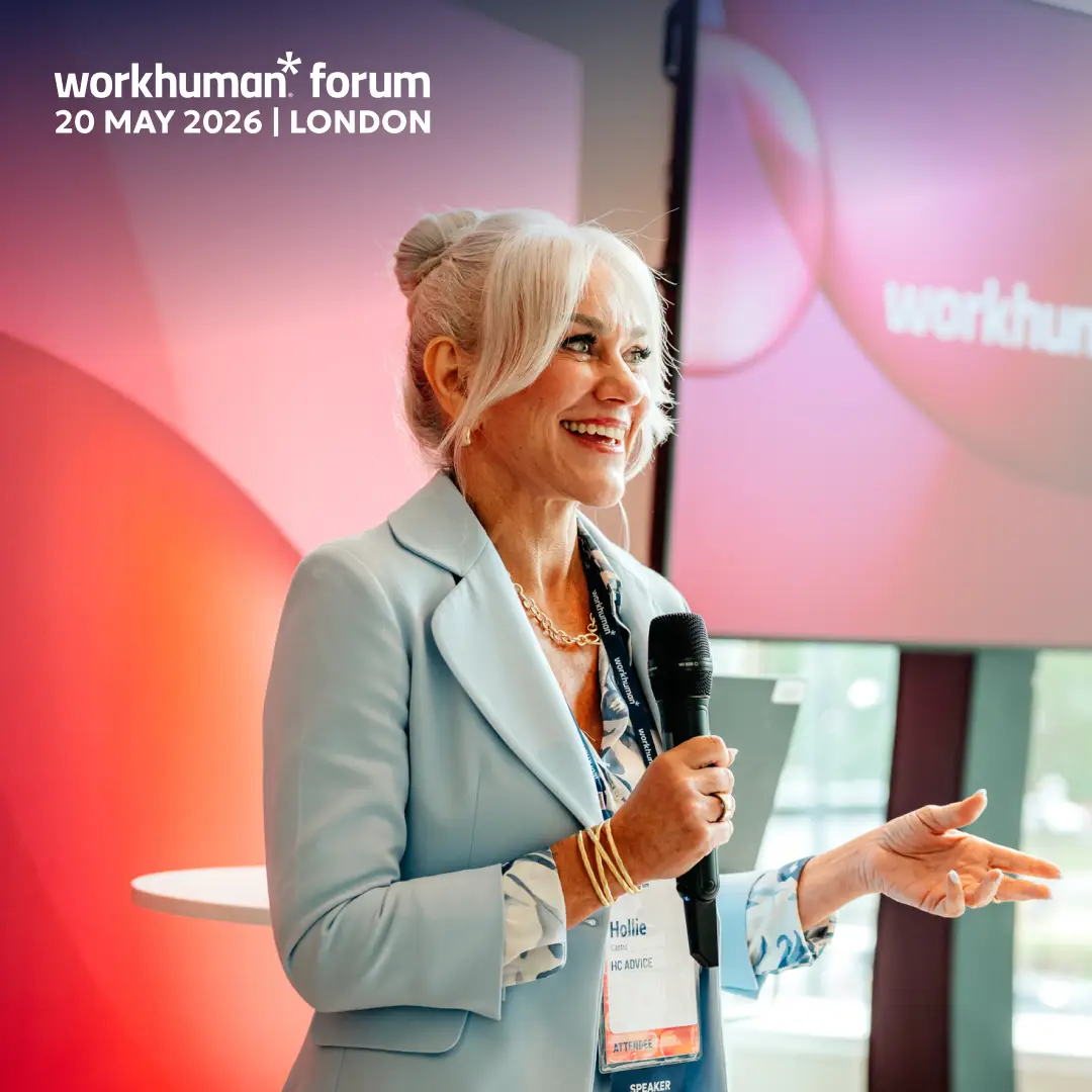 A woman with blonde hair styled in a bun is smiling while speaking into a microphone at the Workhuman Forum event in London on May 20, 2026. She is wearing a light blue blazer over a floral blouse and has a name tag that reads "Hollie" along with "HC ADVICE" and "SPEAKER." In the background, there are colorful abstract shapes along with the Workhuman logo visible on a screen.