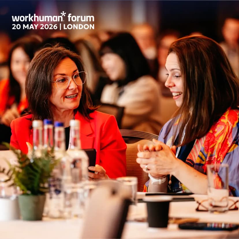 A woman in a red blazer is engaging in a lively conversation with another woman while seated at a table during the Workhuman forum event. The background includes blurred outlines of other attendees, with items like water bottles and plants visible on the table. Overlay text indicates the event title and date: "workhuman* forum 20 MAY 2026 | LONDON."