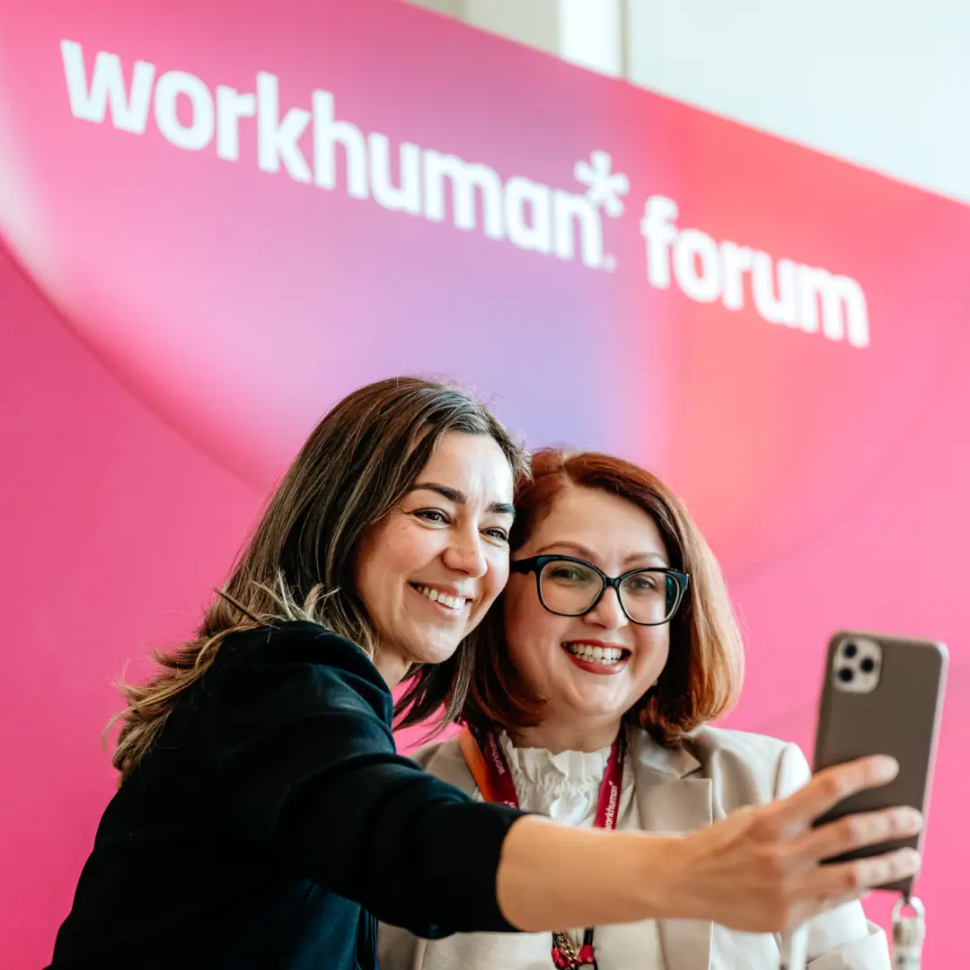 Two smiling women take a selfie in front of a vibrant pink background with the text "workhuman* forum" prominently displayed. One woman has long brown hair and wears a black jacket, while the other has short reddish-brown hair, glasses, and a light-colored blouse, showcasing a joyful moment at the event.