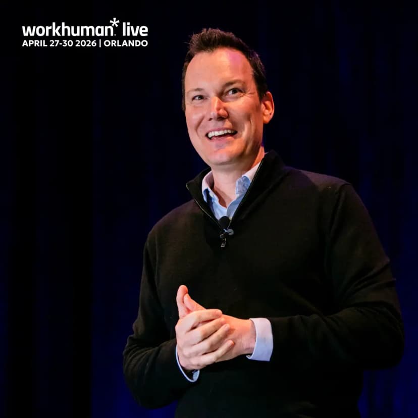 A smiling speaker stands on stage, dressed in a dark sweater over a collared shirt, with hands together as if engaged in conversation. The background is dark, creating a spotlight effect, and there is text at the top left corner that reads "workhuman* live APRIL 27-30 2026 | ORLANDO."