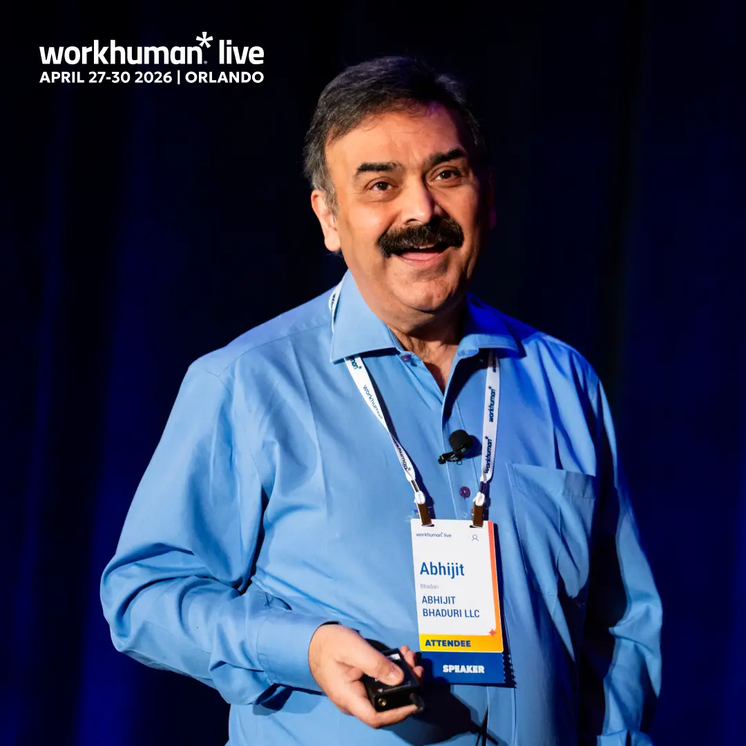 A smiling man stands in front of a dark backdrop, wearing a blue shirt and holding a microphone. He has a name tag that reads "Abhijit Bhaduri LLC" with the titles "ATTENDEE" and "SPEAKER." The image features the text "workhuman live APRIL 27-30 2026 | ORLANDO" at the top.