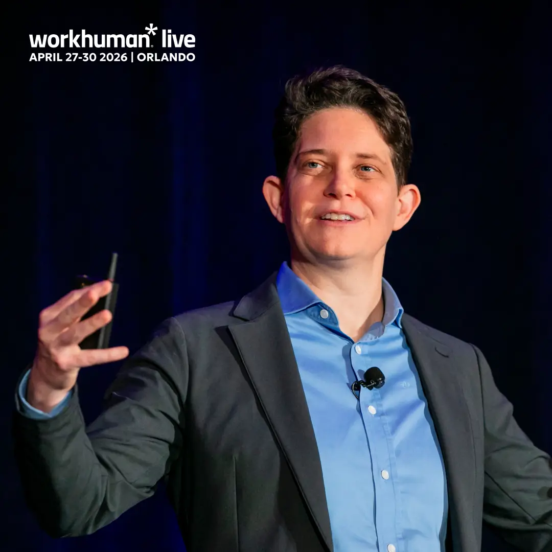 A speaker with short hair and a black blazer is presenting on stage, gesturing with one hand while holding a microphone in the other. The background is dark, highlighting the speaker and a large display reads "workhuman* live APRIL 27-30 2026 | ORLANDO" at the top left of the image.
