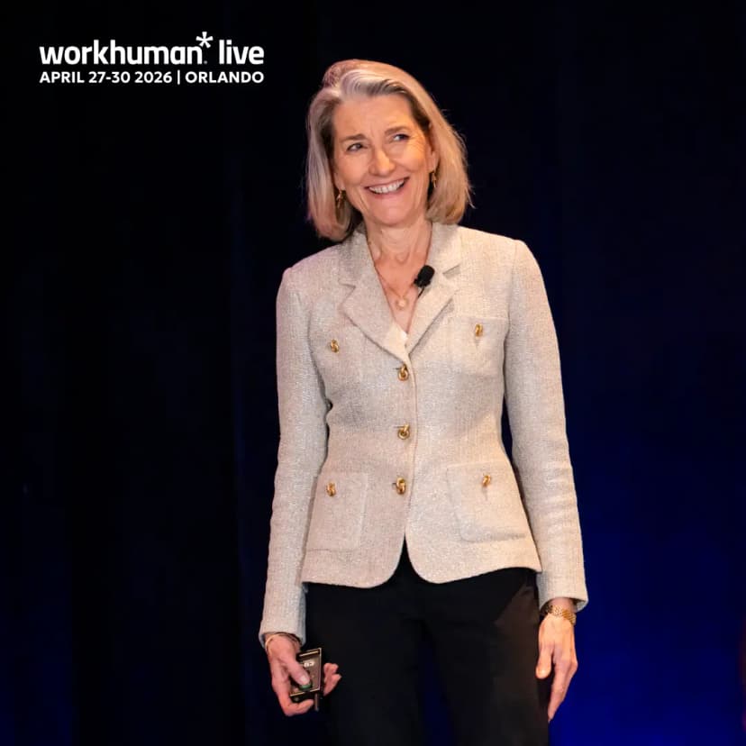 A smiling woman stands on stage, dressed in a light grey jacket with gold buttons and black pants. She holds a small device in her hand and appears to be engaging with an audience. The background is dark, highlighting her figure. Text at the top reads "workhuman live APRIL 27-30 2026 | ORLANDO."