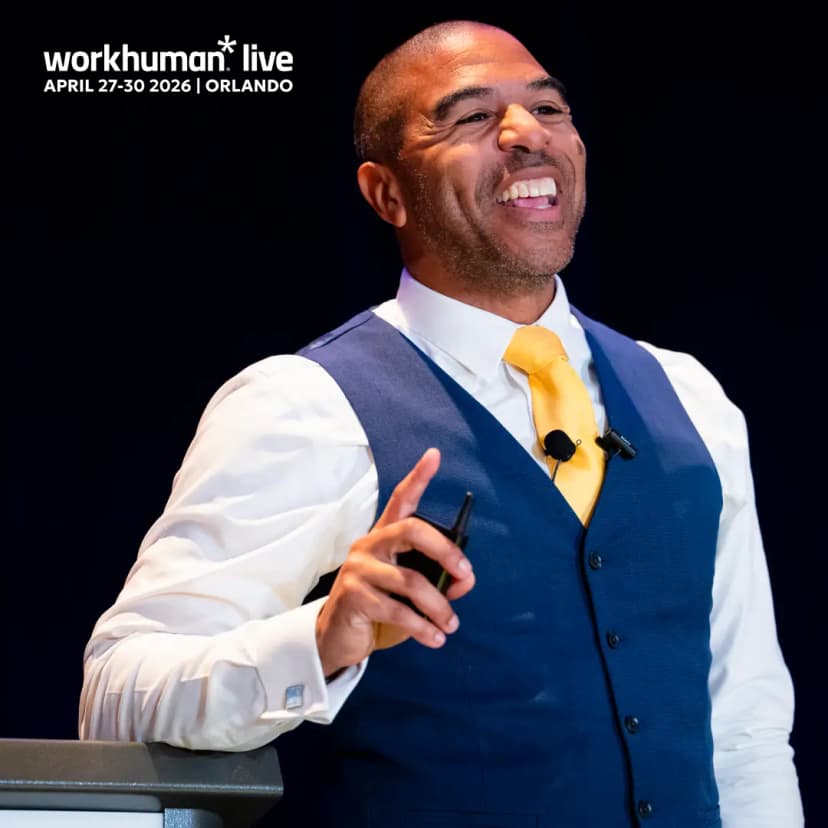 A smiling speaker wearing a blue vest and a yellow tie gestures with one hand while holding a pen in the other. He is engaging with an audience, with a backdrop displaying "workhuman live" and the dates April 27-30, 2026, in Orlando.