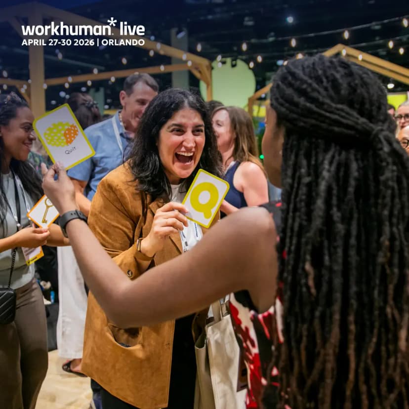 A lively conference scene featuring excited attendees engaging with each other. One woman in the foreground, smiling broadly, is holding cards displaying the words "Quit" and the letter "Q." Other participants are also visible in the background, enjoying the interactive atmosphere at the Workhuman Live event, which is scheduled for April 27-30, 2026, in Orlando. The event branding is prominent in the upper corner of the image.
