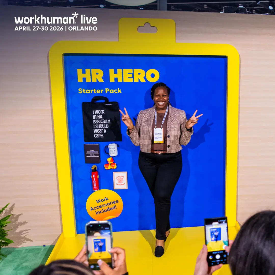 A smiling woman is posing in front of a colorful display at the Workhuman Live event. The display features the text "HR HERO Starter Pack" prominently, alongside various items including a black tote bag with the slogan "I work in HR. Basically, I SHOULD wear a cape," and other accessories. Several attendees can be seen taking photos of her with their smartphones, creating a lively atmosphere. The overall scene reflects a celebration of human resources professionals and their contributions.
