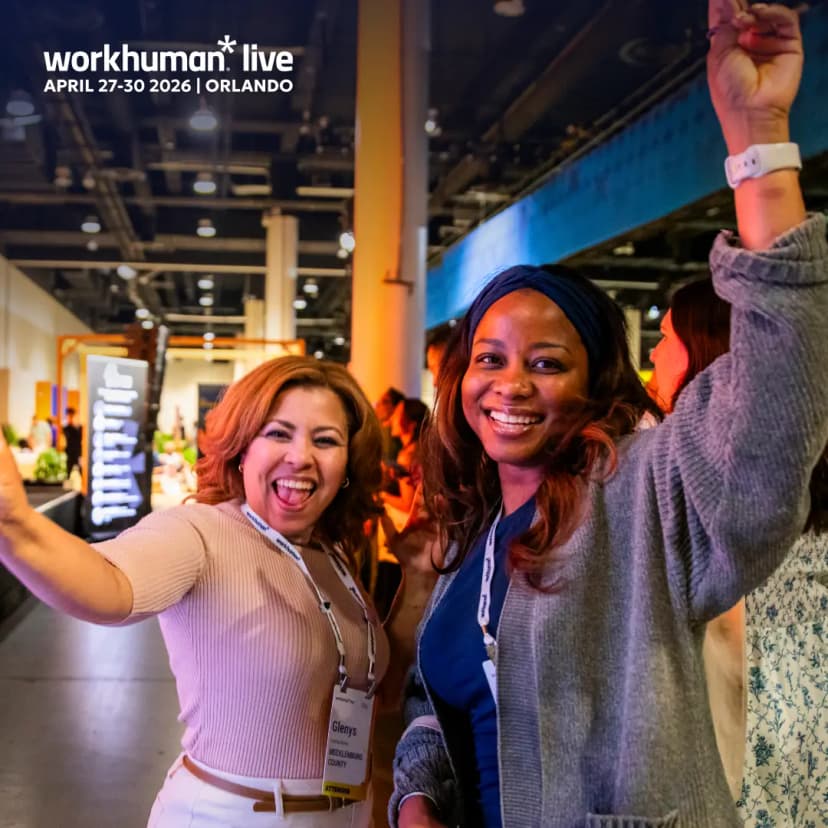 Two women are joyfully posing for a photo at a Workhuman Live event in Orlando, showcasing smiles and excitement. One woman has reddish-brown hair and is wearing a light beige top, while the other has dark hair styled with a headband and is dressed in a blue top. Both have name tags visible. The atmosphere is lively, with colorful lighting and event branding in the background, emphasizing community and celebration within the professional setting.