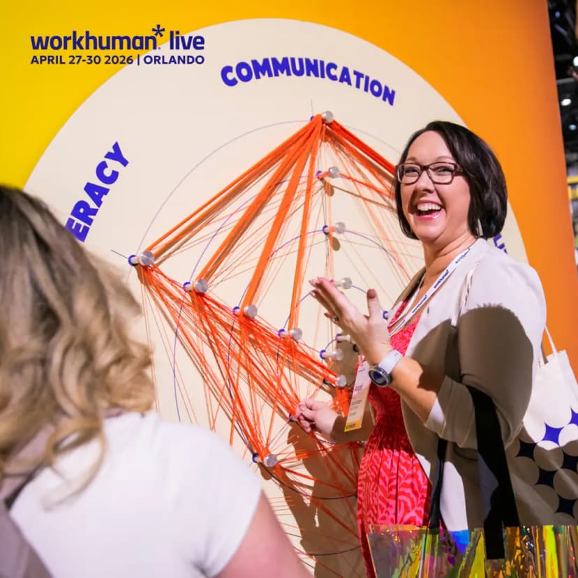 A woman smiling and engaged in interaction stands beside a visual display at the Workhuman Live event. The display features interconnected strings and nodes illustrating themes of "Communication" and "Literacy," with the event dates, April 27-30, 2026, and location, Orlando, prominently displayed at the top. She gestures animatedly while explaining the display, conveying a sense of enthusiasm and participation in the event.
