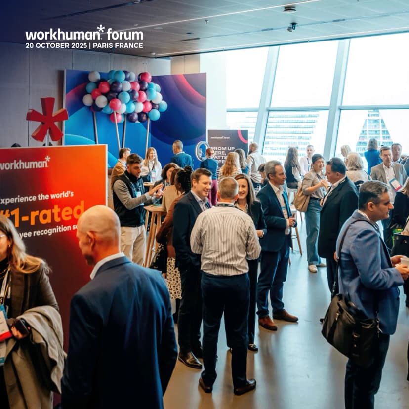 A busy conference event featuring the Workhuman Forum, taking place on October 20, 2025, in Paris, France. Attendees are engaged in conversations while gathered around tables. Prominent in the scene is a large display featuring the Workhuman logo and a message indicating it is the world's number one rated employee recognition software. A colorful backdrop of balloons adds to the ambiance.