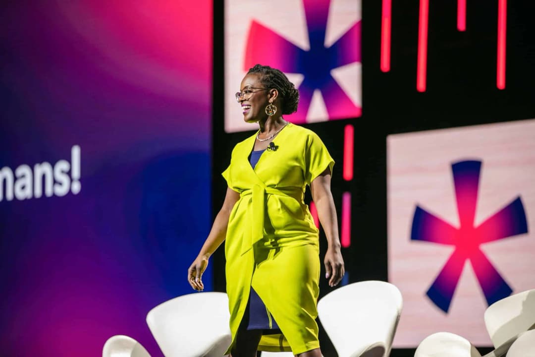 A woman wearing a bright yellow dress stands on stage with a smile, addressing an audience. She is positioned in front of a colorful backdrop that features the word 'Humans!' along with graphic elements, including large stylized icons and dynamic lighting. Two white chairs are set on the stage beside her.