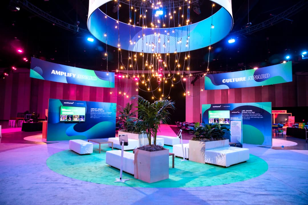 A vibrant display at the Workhuman Live conference showcasing the Amplify Award and Culture Award. The area features a large, illuminated overhead structure with hanging lights and colorful banners. In the foreground, there are seating arrangements with white couches and potted plants. Prominent blue and green signage highlights the awards, with text emphasizing values such as 'creating ripple effects of positive culture' and ensuring safety in the workplace. The overall setting reflects an energetic and engaging atmosphere for attendees.