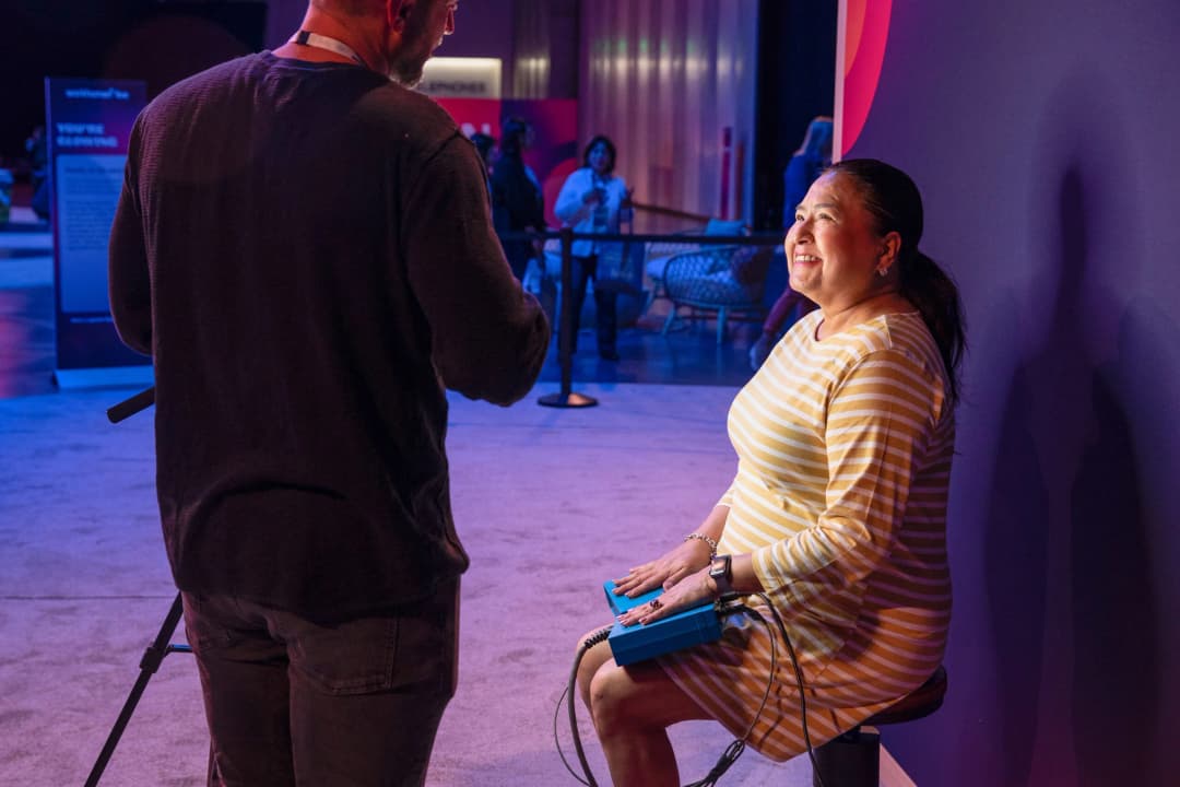 A woman sitting on a stool smiles as she interacts with a man standing in front of her. She wears a striped dress and has devices connected to her hands. The setting appears to be a conference or event, with blurred attendees visible in the background and colorful lighting creating a vibrant atmosphere.