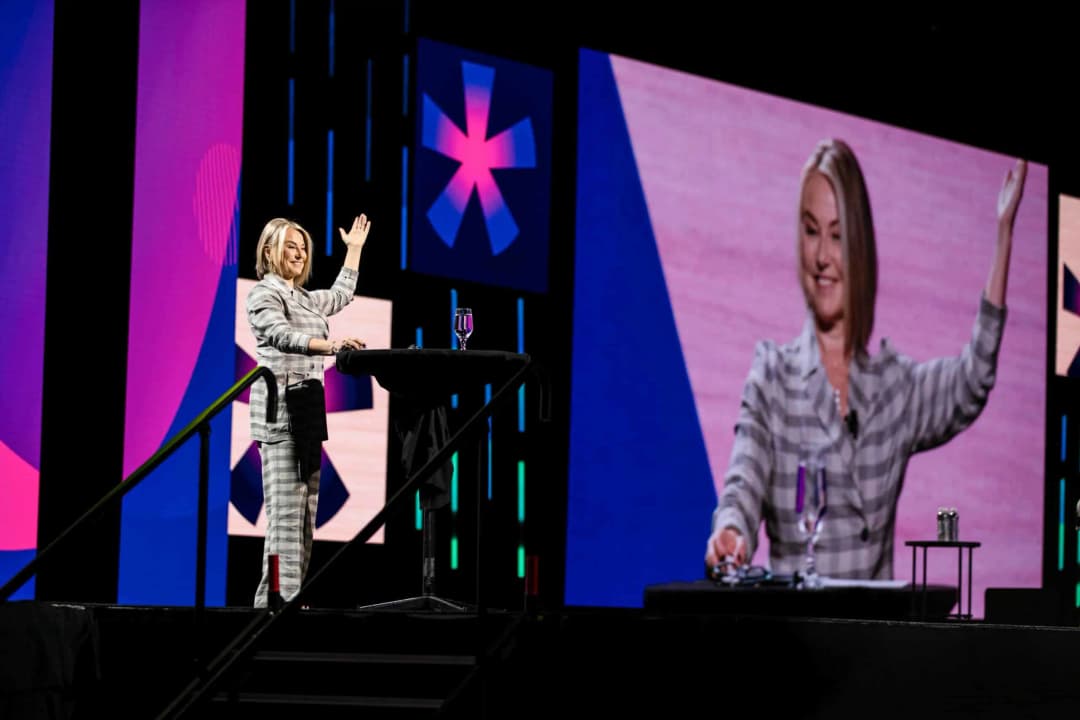 A speaker on stage is waving, engaging the audience at a Workhuman event. She is wearing a grey and white checkered outfit and standing next to a table with a glass of water and some items. Two large display screens show her image and vibrant background colors in shades of pink and blue.