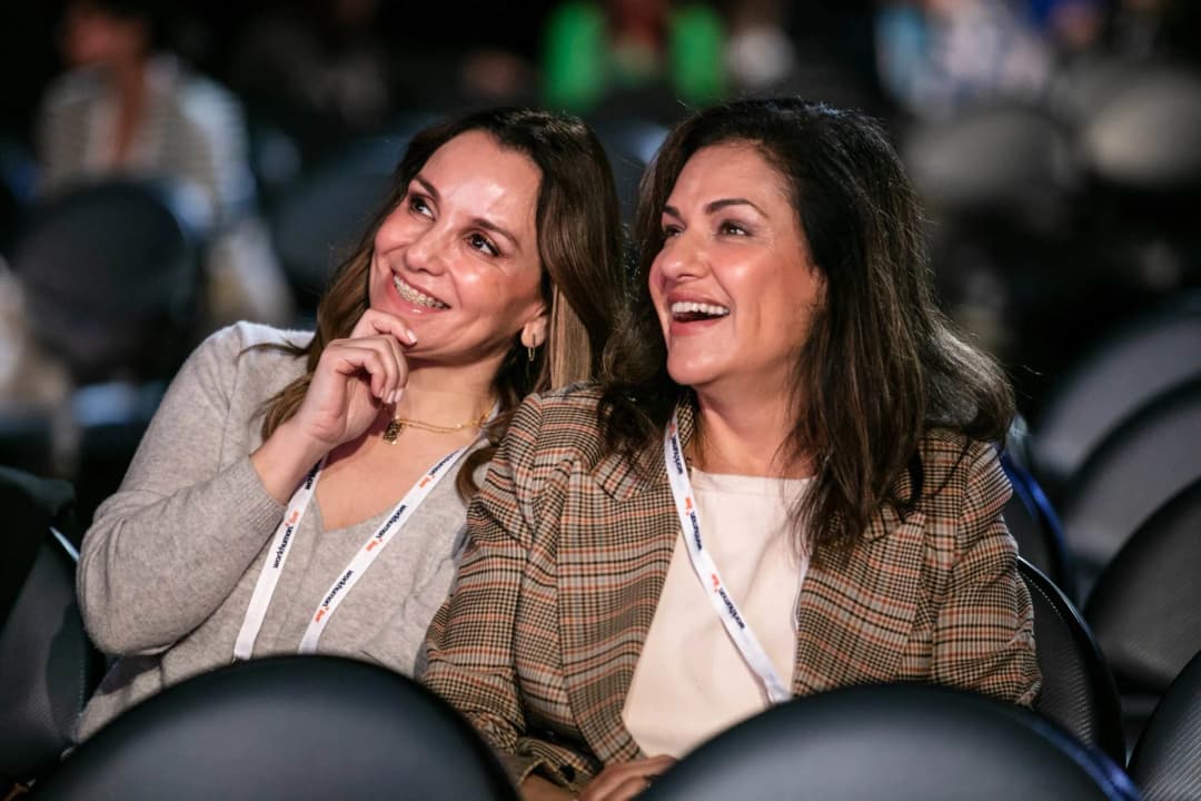 Two women are seated in an audience, smiling and engaged in conversation during the Workhuman Live conference. Both are wearing lanyards and are dressed casually. The background features blurred outlines of other attendees and empty chairs.