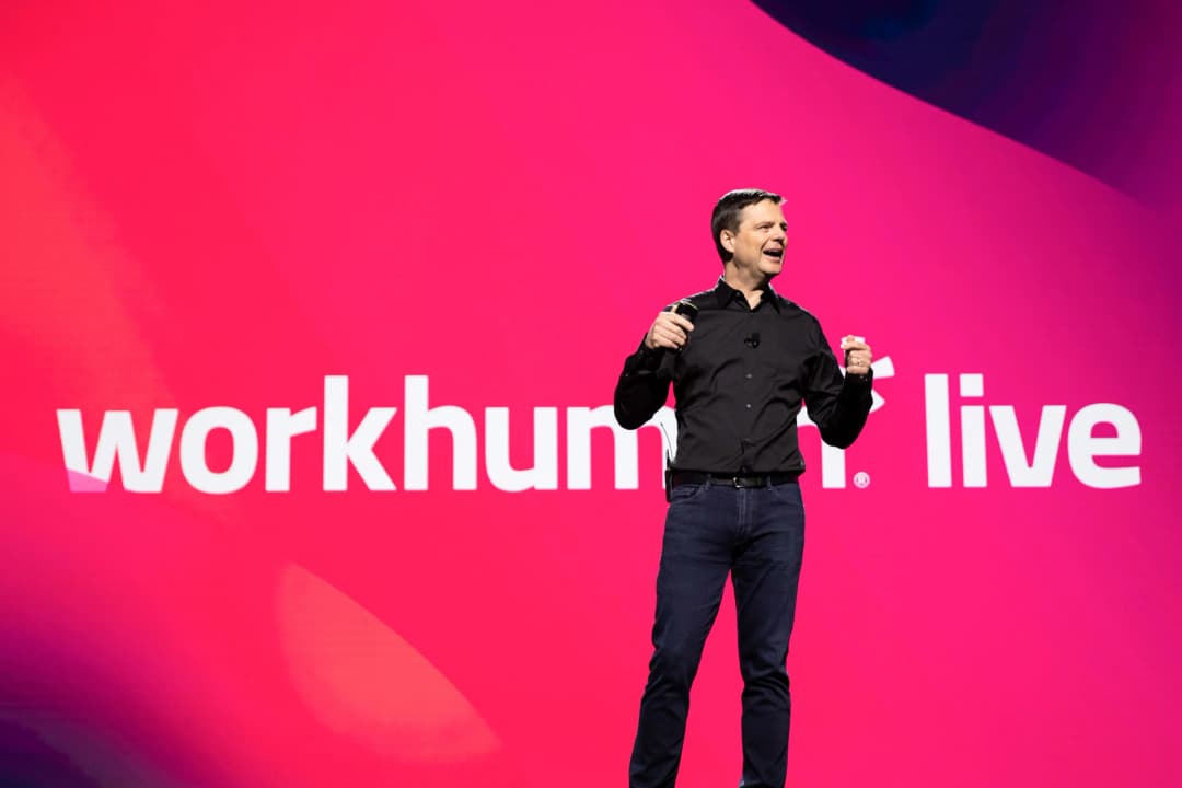 A speaker is presenting on stage at a Workhuman Live event, standing confidently with a microphone in hand. Behind him is a large, vibrant pink display featuring the text 'workhuman. live.' The atmosphere appears engaging and energetic, suggesting an interactive or informative session.