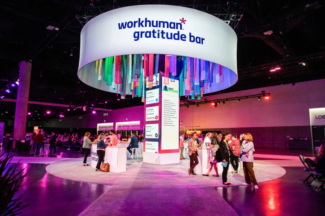 A vibrant Workhuman Gratitude Bar setup at a conference featuring colorful ribbons hanging from a circular sign that reads 'workhuman gratitude bar.' Attendees engage in conversation and activities around several high-top tables and displays, with sections showing various initiatives like 'Project New Village' focused on community well-being. Surrounding areas are well-lit with a mix of purple and warm lights, and signs indicating amenities such as restrooms are visible in the background.