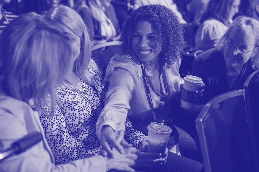 A group of four women engage in conversation, smiling and interacting warmly with each other. One woman is extending her hand to another, possibly for a handshake or gesture of connection. They are seated in a conference venue with other attendees visible in the background. Some women are holding beverages, creating a casual and friendly atmosphere. The image has a blue tone applied, giving it a unified look.