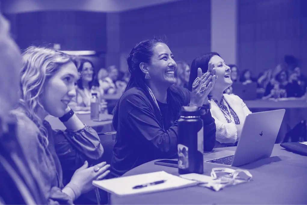Attendees at a conference are engaged and smiling as they enjoy a presentation. One woman is clapping enthusiastically, while others look on with expressions of enjoyment. A laptop and water bottle are on the table in front of them, indicating an interactive and collaborative environment. The setting appears lively and supportive, fostering a sense of community among the participants.