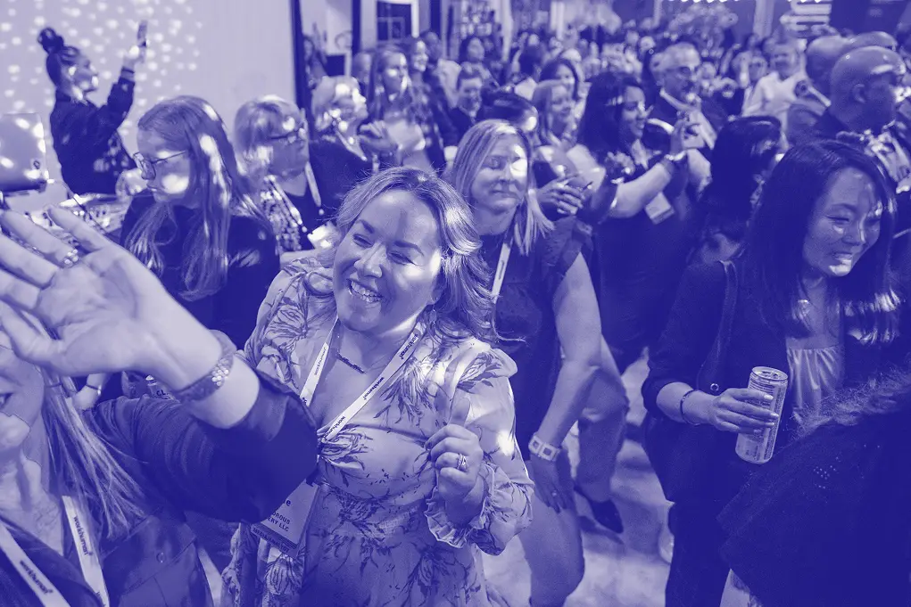 A lively scene at a event with a crowd of attendees enjoying themselves. Several women are prominently seen interacting and smiling, with one woman in the foreground joyfully dancing and engaging with another attendee. The atmosphere appears festive, highlighted by soft, ambient lighting. Party-like decor can be seen in the background, contributing to the celebratory vibe of the gathering.