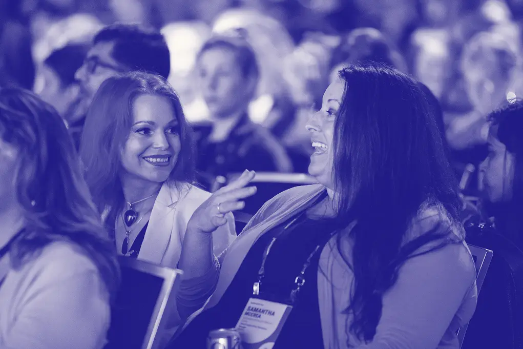 Two women are engaged in a lively conversation at an event, both smiling and appearing to be in good spirits. The setting is filled with attendees in the background, indicating a social atmosphere. One woman, with long hair and a necklace, gestures as she speaks, while the other, with darker hair, laughs and responds enthusiastically. Both women seem to be enjoying the moment, reflecting a sense of camaraderie and connection.