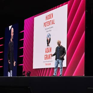 A speaker, Adam Grant, stands on a stage presenting at an event. Behind him is a large screen displaying the title "HIDDEN POTENTIAL" along with his name. The stage features bold pink and purple lighting, creating a vibrant backdrop for his talk.