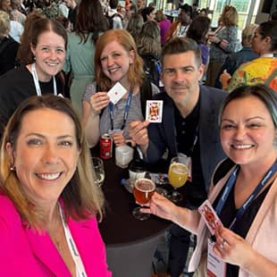 A group selfie featuring five people smiling together at an event, with a bustling crowd in the background. The individuals are holding drinks and some have playing cards, showcasing a vibrant and engaging atmosphere. They appear to be enjoying the moment in a social setting, likely a conference or gathering.