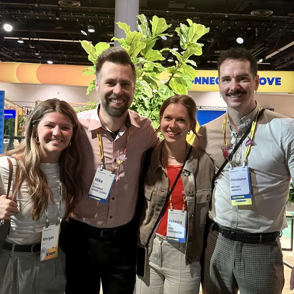 A group of four individuals is smiling together at an event, standing in front of a plant backdrop. Each person is wearing a name tag, indicating their names and affiliations: Morgan (staff), Mike (Resort Casino), Rebecca (AGCO Corporation), and Christopher (British Airways). The setting reflects a lively atmosphere typical of a professional conference.