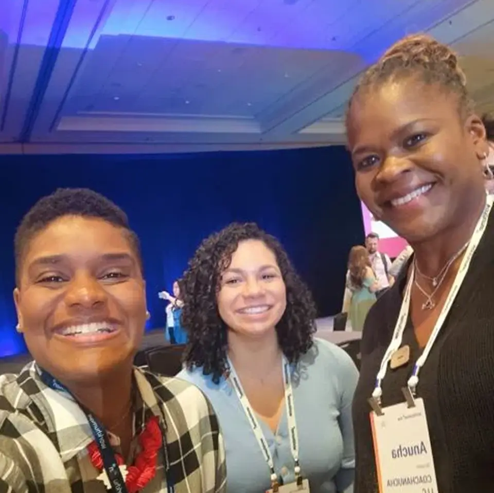 A group selfie of three smiling women at a conference, with a dimly lit stage in the background. Each woman wears a lanyard with their name tags visible. The woman on the left has short hair and is wearing a checked shirt, the middle woman has curly hair and is wearing a light sweater, and the woman on the right has long hair tied back, wearing a black sweater. The atmosphere suggests a friendly and engaging conference environment.